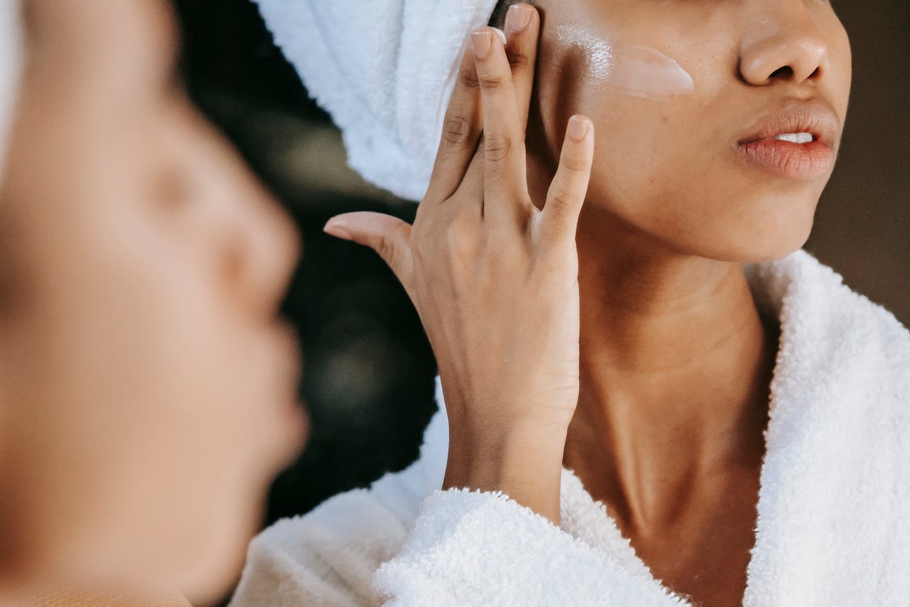 Home Crop unrecognizable young ethnic female in terry robe applying moisturizing cream on cheek while reflecting in mirror